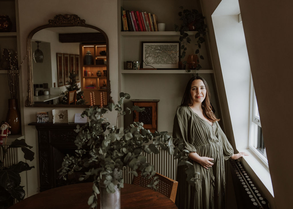 Expectant mother in green flowing dress by window during in-home maternity photography session in London