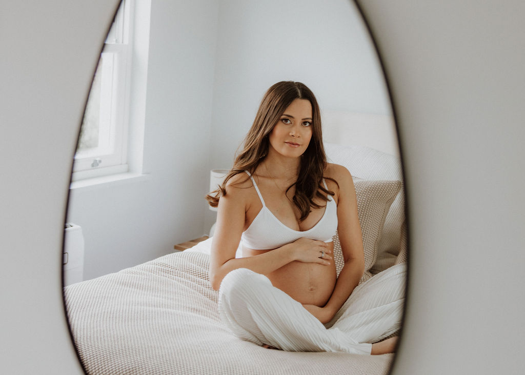 Pregnant mother gazing into a mirror and softly touching her baby bump during a luxury in-home maternity photography session in London.