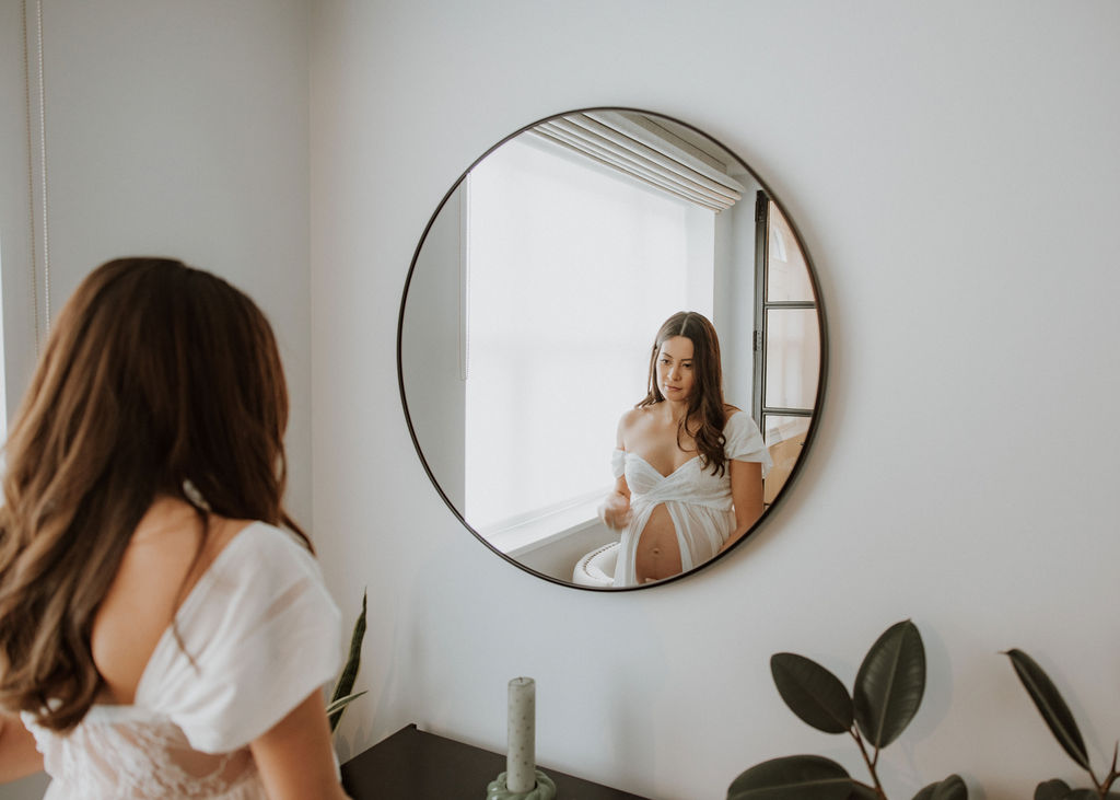 Expectant mother gazing at reflection in bedroom mirror during in-home maternity photography session in London